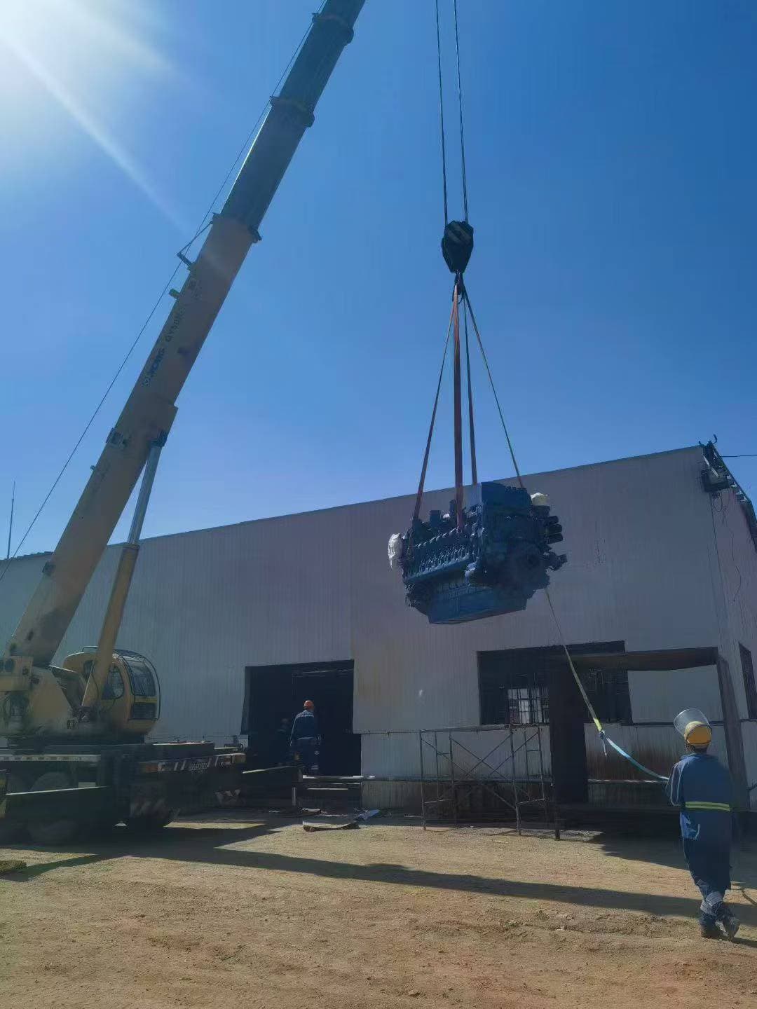 A crane lifting the engine block into position above the workshop doors while two workers guide the slings