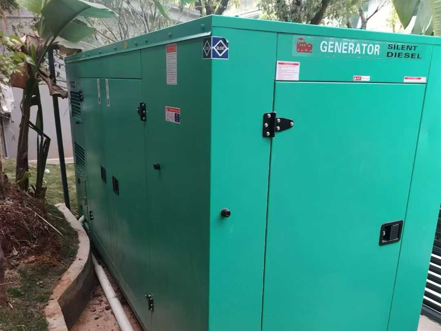 A green 300 kW silent canopy set standing on a grass verge among palm trees beside a hotel building